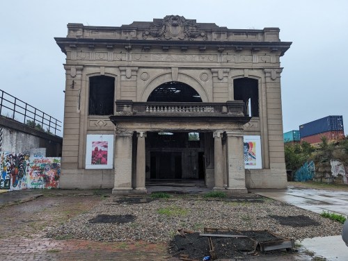 Abandoned building with ornate architecture, partially collapsed, surrounded by graffiti and debris on a cloudy day.