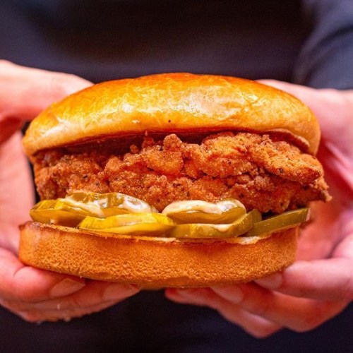 A close-up of a crispy fried chicken sandwich with pickles on a soft bun, held by two hands.