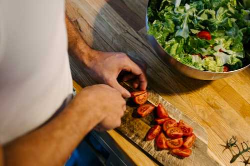 A person slices tomatoes on a wooden cutting board next to a bowl of salad.