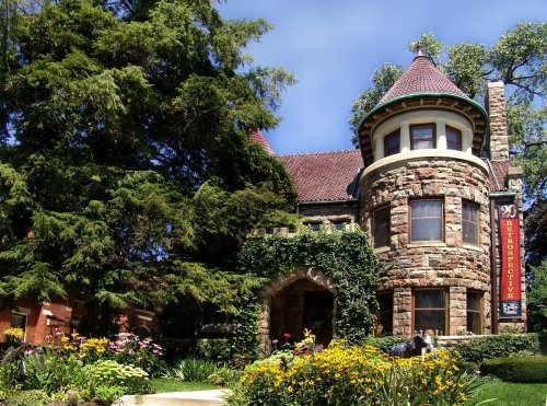 A stone building with a turret, surrounded by colorful flowers and greenery, under a clear blue sky.