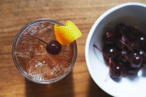 A refreshing cocktail with ice, a cherry, and a twist of orange peel, next to a bowl of cherries on a wooden surface.