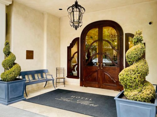 Entrance of Hotel Les Tres featuring elegant wooden doors, topiary plants, and a bench.
