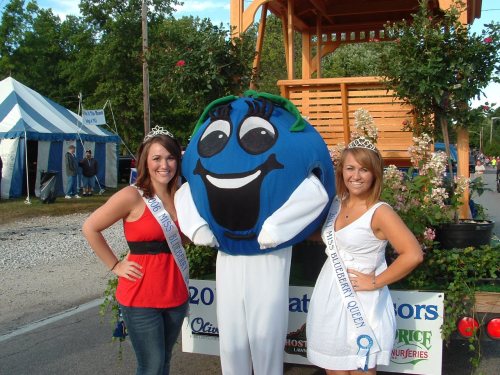 Two women in tiaras pose with a large blueberry mascot at a parade, surrounded by flowers and a festive backdrop.
