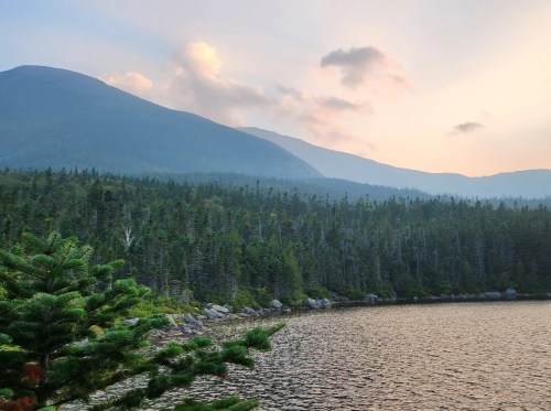 A serene lake surrounded by dense forest and mountains under a soft, cloudy sky at sunset.