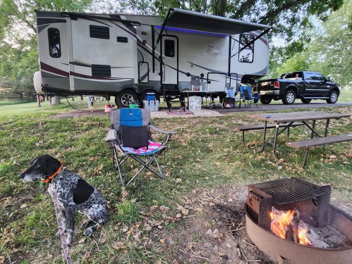 A camping scene with a parked RV, a dog sitting nearby, a fire pit, and outdoor chairs under a canopy.