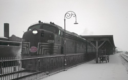 A vintage train at a snowy station, with a lamppost and a cart nearby, evoking a nostalgic winter scene.