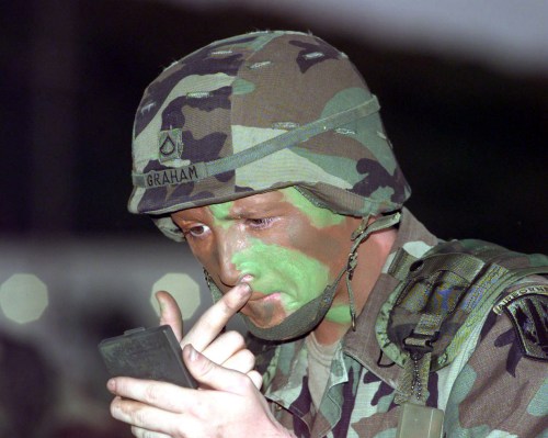 A soldier in camouflage uniform and helmet applies face paint while looking at a small mirror.