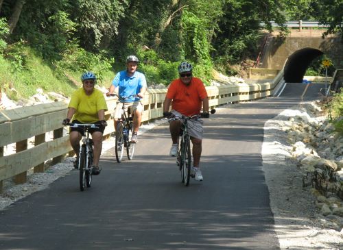 Three cyclists ride on a paved path surrounded by greenery, approaching a tunnel in the background.