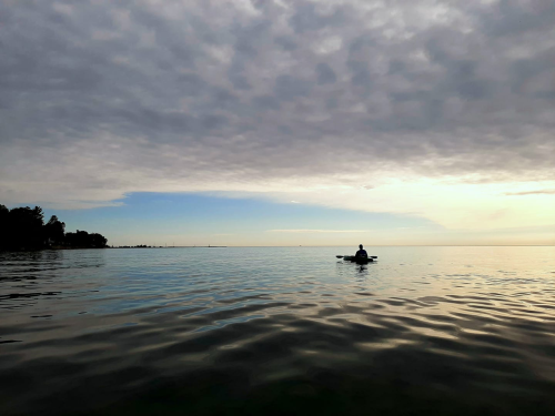 A lone kayaker paddles on calm waters under a cloudy sky, with a distant shoreline visible.