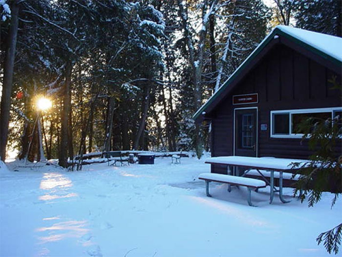 A cozy cabin in a snowy landscape, surrounded by trees, with picnic tables and a sunset in the background.