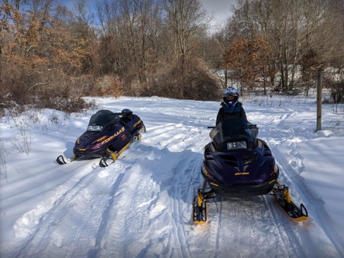 Two snowmobiles parked on a snowy trail, with a person wearing a helmet standing next to one of them.