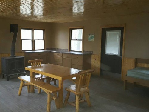 A rustic interior of a cabin featuring a wooden table, chairs, a stove, and large windows.