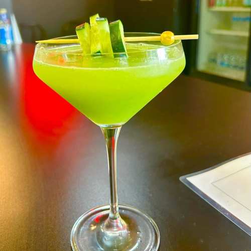 A vibrant green cocktail garnished with cucumber slices, served in a stylish martini glass on a dark bar surface.