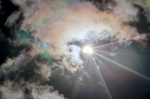Bright sun partially obscured by colorful, swirling clouds against a dark sky.