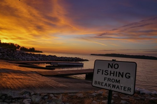 A sunset over a calm lake with a sign reading "No Fishing from Breakwater" in the foreground.