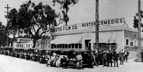 Historic black-and-white photo of a film company building with vintage cars and people posing outside.