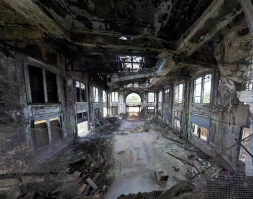 An abandoned, decaying interior of a large building with broken windows and debris scattered on the floor.