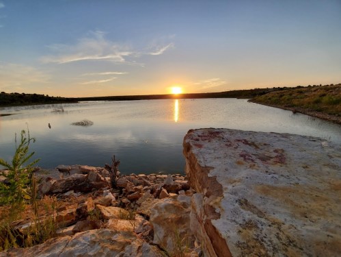 Sunset over a calm lake, with rocky shoreline and gentle hills in the background under a clear sky.