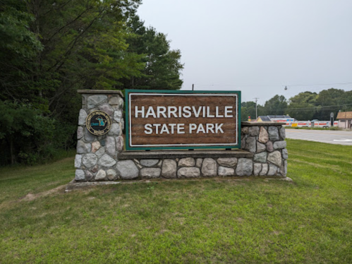Sign for Harrisville State Park, featuring a stone base and green trim, surrounded by grass and trees.