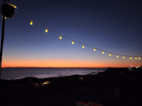 A serene sunset over the ocean, with string lights glowing against a twilight sky.