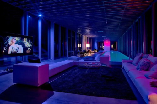 Modern lounge with a pool, illuminated in blue and pink lights, featuring a large TV and stylish furniture.