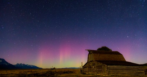 A rustic barn under a starry sky with colorful auroras illuminating the horizon.