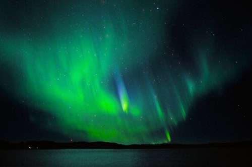 Vibrant green and blue auroras dance across a starry night sky over a calm lake.