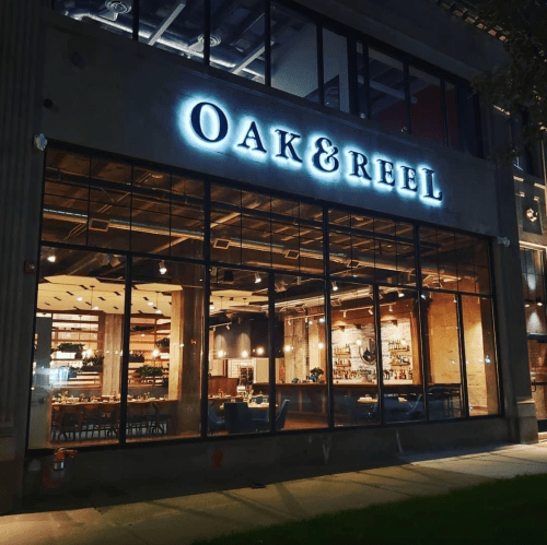 Exterior view of Oak & Reel restaurant at night, featuring large windows and illuminated signage.