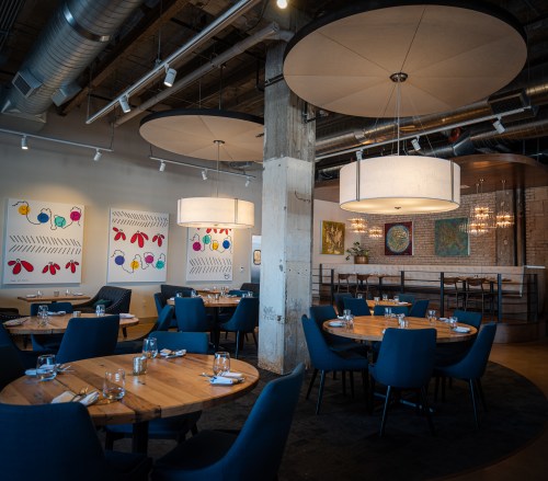 A modern restaurant interior featuring round tables, blue chairs, and colorful artwork on the walls.