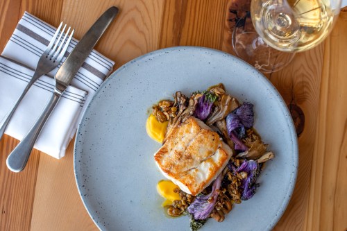 A plated dish featuring seared fish atop colorful vegetables and grains, accompanied by a glass of white wine.