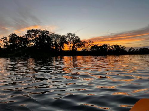 A serene sunset over a calm lake, with silhouetted trees reflecting on the water's surface.