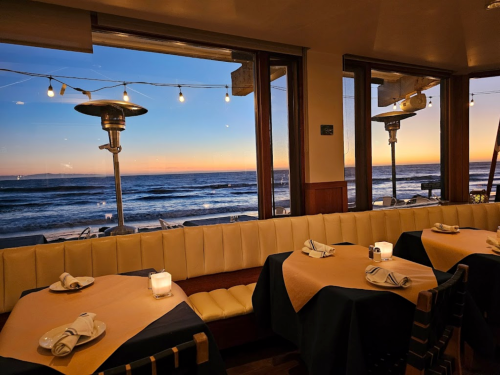 Cozy restaurant interior with tables set for dinner, overlooking a sunset-lit beach and ocean through large windows.