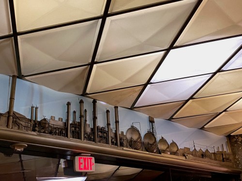 A decorative wall mural featuring industrial structures and pipes, set against a geometric ceiling. An exit sign is visible.