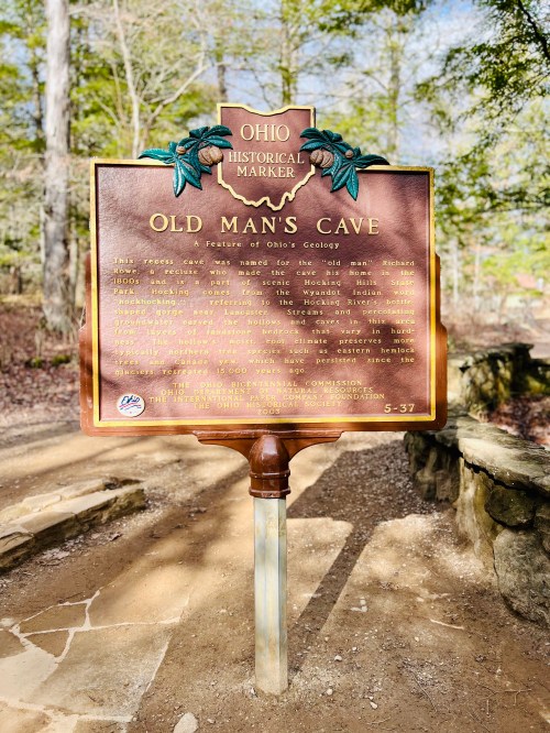Historical marker for Old Man's Cave, detailing its significance in Ohio's geology, surrounded by trees.