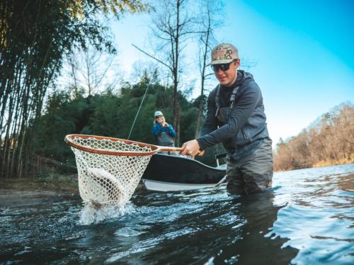 A fisherman in waders nets a fish in a river, while another person watches from a boat in the background.