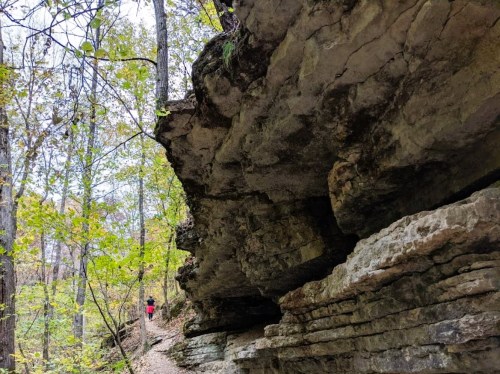 A rocky cliffside along a forest trail, with a person in red walking in the distance among trees and autumn foliage.