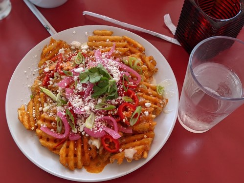 A plate of loaded waffle fries topped with cheese, pickled onions, cilantro, and sliced peppers, next to a glass of water.