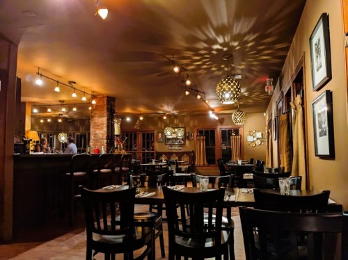 Cozy restaurant interior with wooden tables, black chairs, warm lighting, and decorative wall art.
