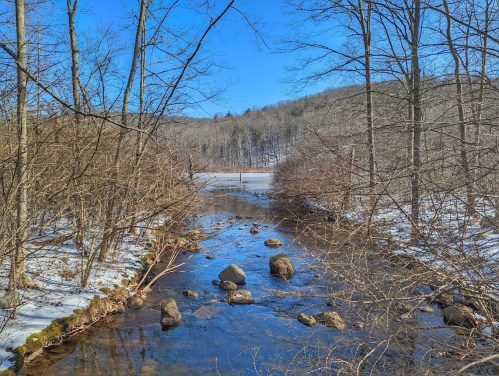 A serene stream flows through a snowy landscape, surrounded by bare trees and distant hills under a clear blue sky.