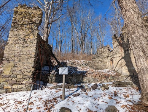 Ruins of a stone structure surrounded by trees and snow, with a sign indicating historical significance.