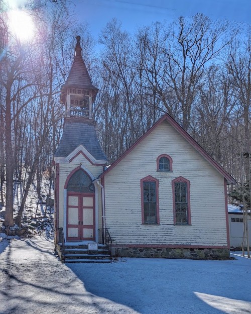 A quaint, snow-covered church with a tall steeple, surrounded by bare trees in a winter landscape.