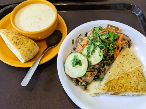 A plate with grilled chicken topped with vegetables, served with garlic bread and a bowl of creamy soup.