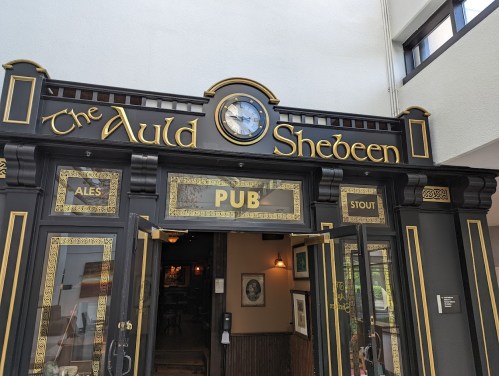 Entrance of "The Auld Shebeen" pub, featuring a clock and decorative signage for ales and stout.