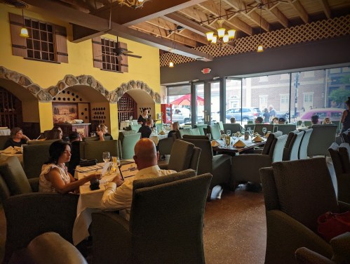 A spacious restaurant interior with green chairs, diners at tables, and large windows letting in natural light.