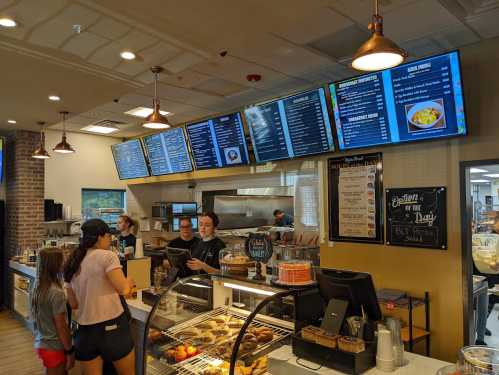 A busy café counter with a menu display, customers ordering, and baked goods in a glass case.