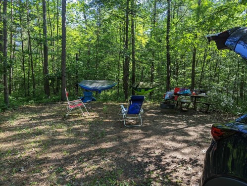 A serene camping spot in a forest with chairs, hammocks, and a picnic table surrounded by trees.