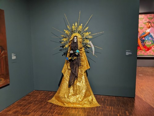 A striking figure in a golden gown with a skull face, adorned with a sunburst halo, displayed against a dark wall.