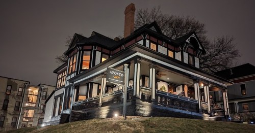 A beautifully lit Victorian-style house at night, featuring a sign that reads "Aposto" and surrounded by greenery.