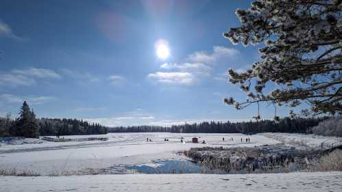 A snowy landscape with people ice fishing on a frozen lake, surrounded by trees and a bright sun in a clear blue sky.