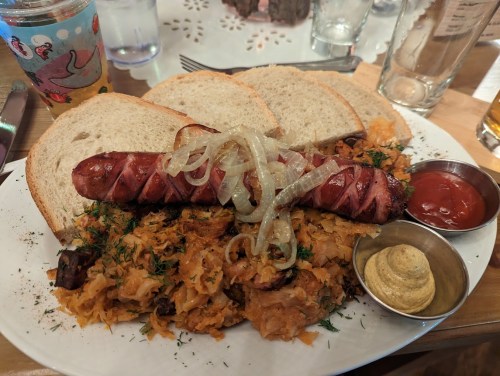 A plate with a sausage topped with onions, served with sauerkraut, mustard, ketchup, and slices of bread.